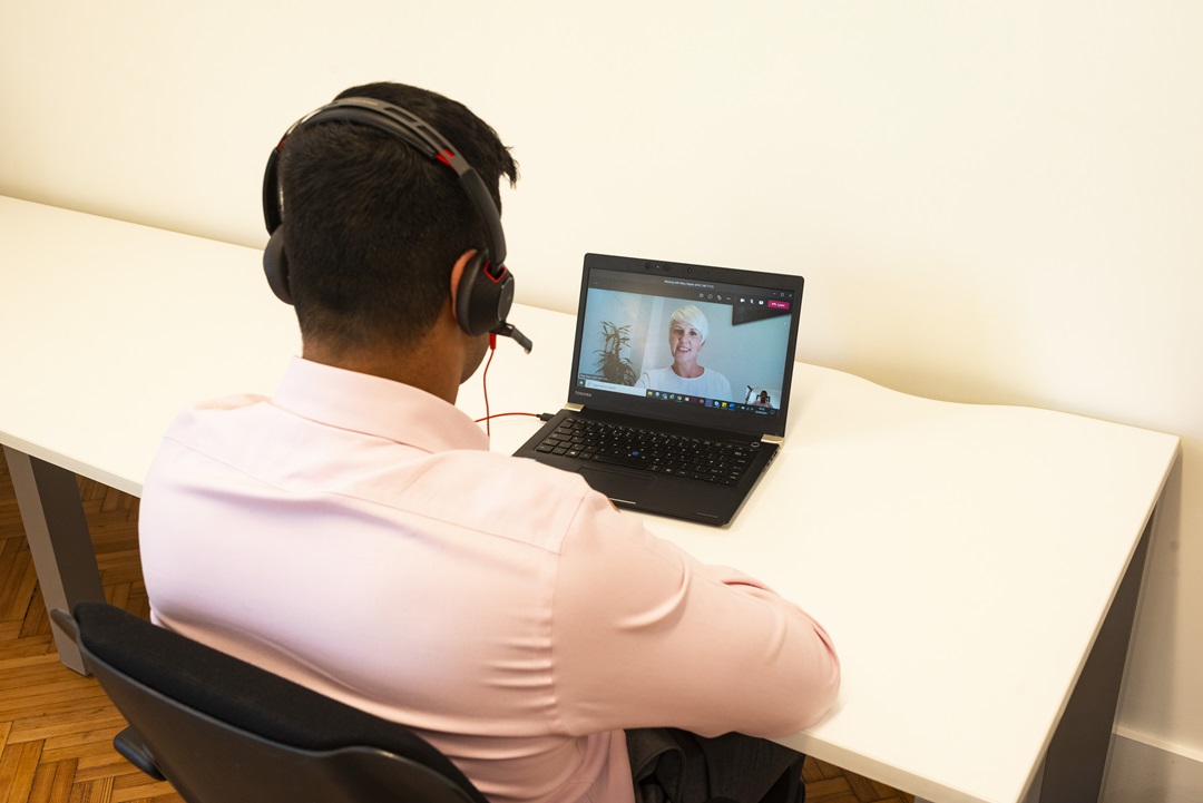 A man sits with his back to the viewer, he's using a headset to speak to a woman in a Teams meeting on the laptop at the table in front of him